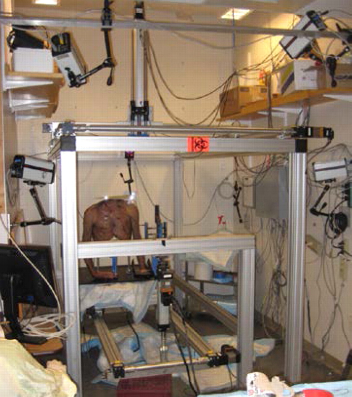 Figs. 2a - 2c 
            Photographs showing a) a saw-bone
skeleton mounted onto the system with the arm fixed to the upper
frame actuator, b) the testing apparatus with the cadaveric torso
and the five-camera setup, with two cameras located on the side,
two above, and one on the back wall behind the torso, and c) the
placement of the five marker clusters on a human torso, each with
three or four individual markers, on the sternum, clavicle, humerus,
scapula and forearm.
          